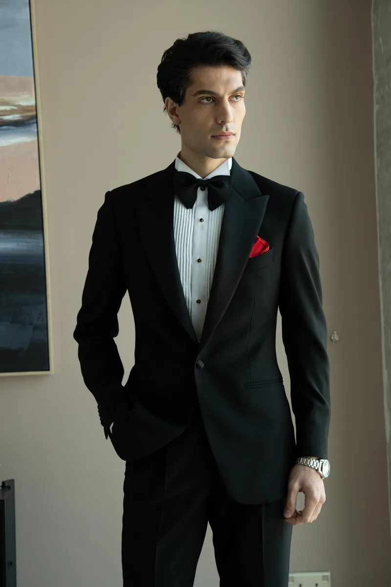 Man adjusting his bespoke black tuxedo jacket beside a window